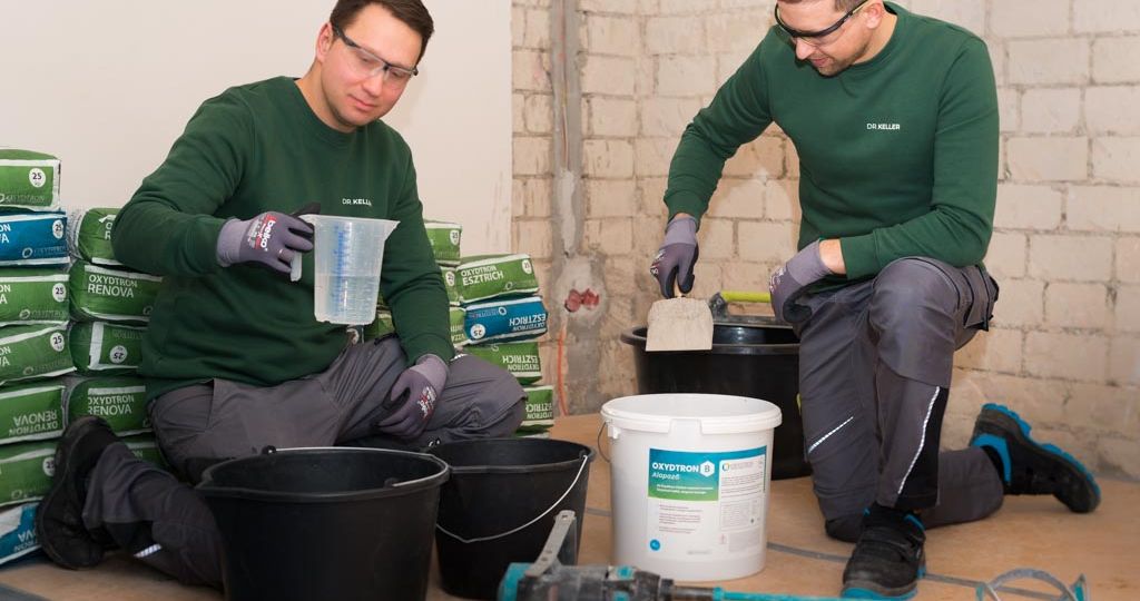 Zwei Handwerker in grünen Pullovern knien beim Mischen von Putz auf dem Boden, umgeben von Oxydtron Baustoffen und Eimern