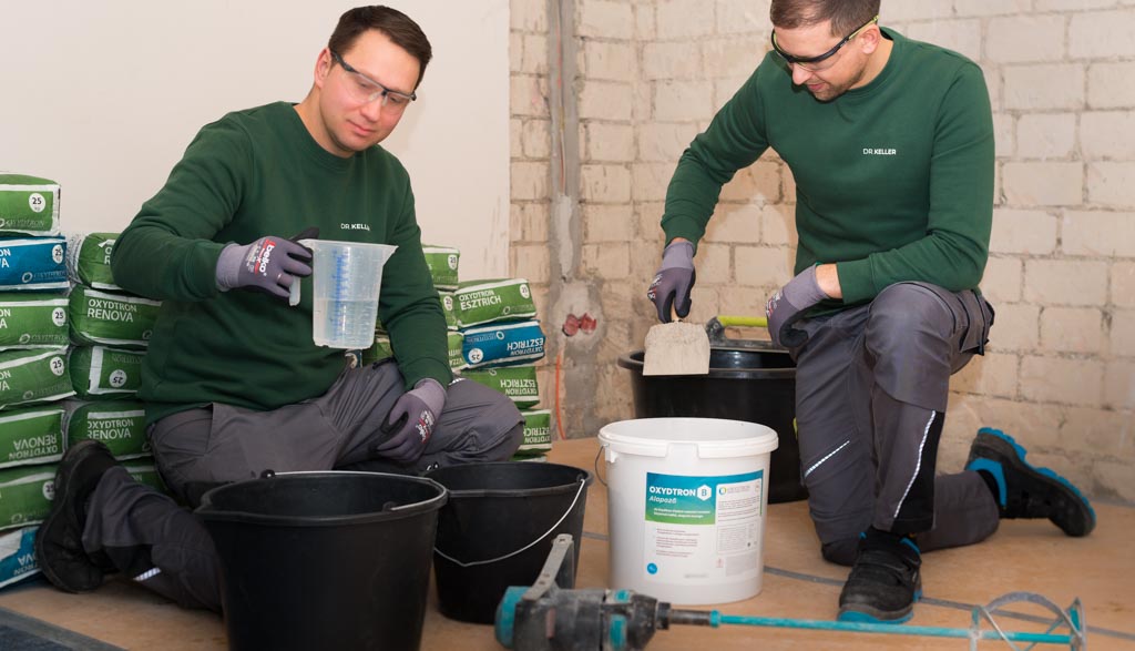 handwerker-beim-mischen-von-putz-auf-dem-boden Zwei Handwerker in grünen Pullovern knien beim Mischen von Putz auf dem Boden, umgeben von Oxydtron Baustoffen und Eimern