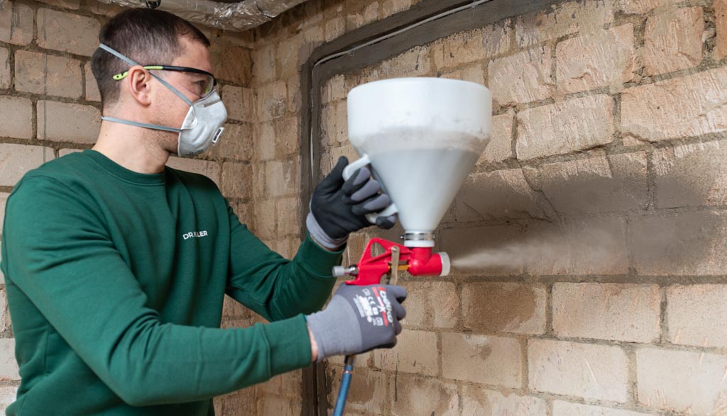 handwerker-sprüht-putz-mit-putztrichter-auf-1 Handwerker mit Atemschutzmaske verwendet einen Putztrichter, um Material an einer Kellerwand aufzusprühen