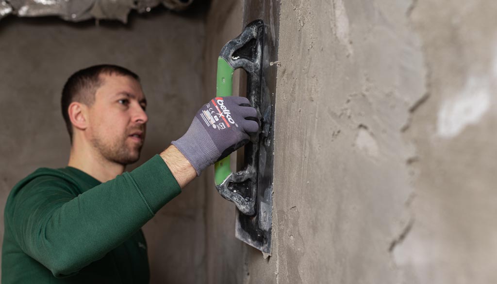 handwerker-mit-spachtel-verteilt-putz-auf-wand Ernster Handwerker in einem grünen Pullover trägt konzentriert Putz auf eine Wand auf, um eine ebene Fläche zu schaffen.