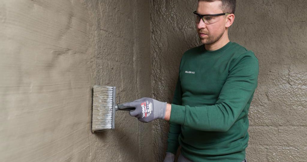 Handwerker in grünem Oberteil trägt mit einer Bürste sorgfältig eine Schutzbeschichtung auf eine Kellerwand auf.