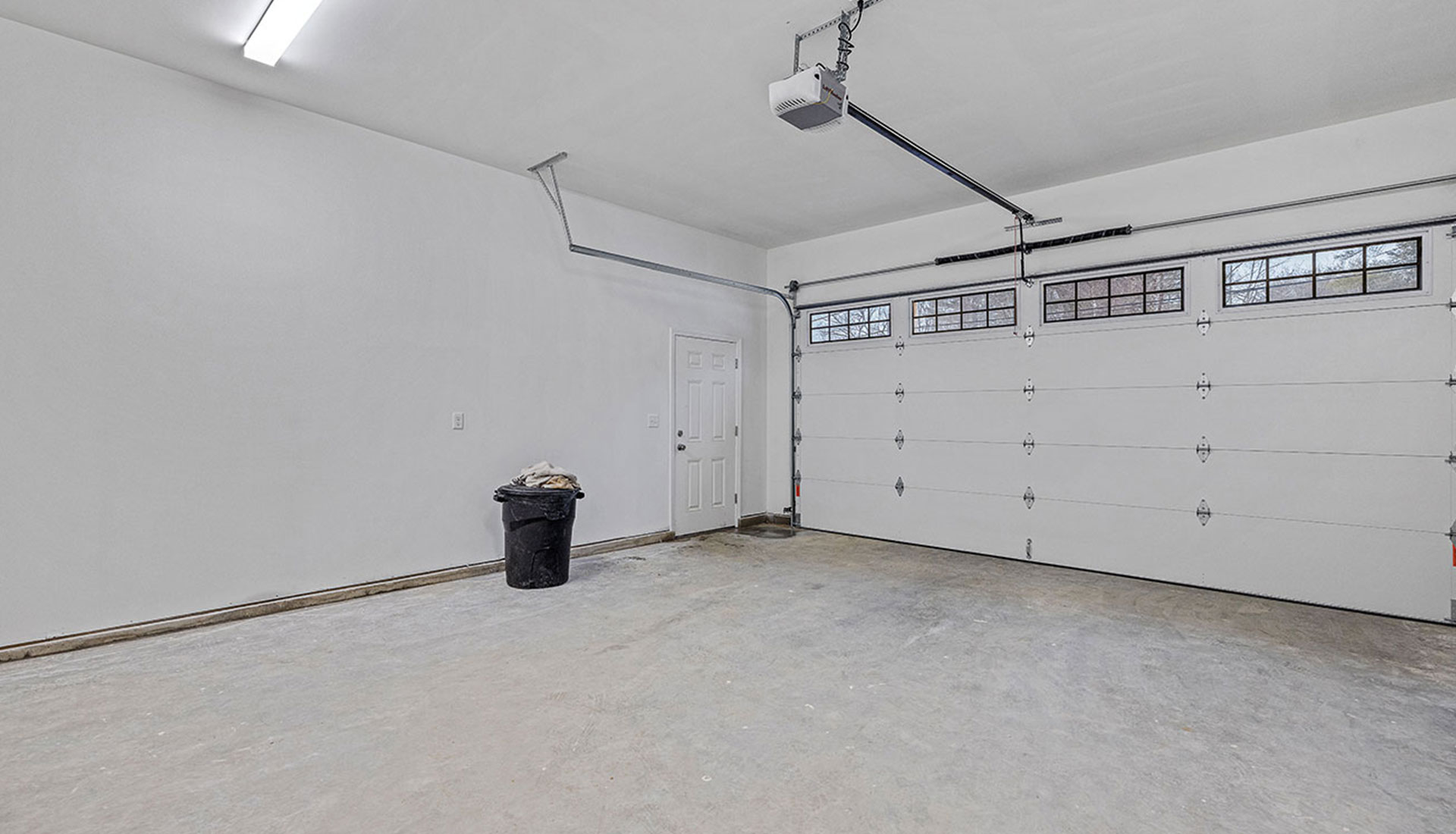 frisch-renovierte-leere-garage Leere frisch renovierte Garage mit weiß gestrichenen Wänden, einer geschlossenen Garagentür und einem einzelnen Abfalleimer.