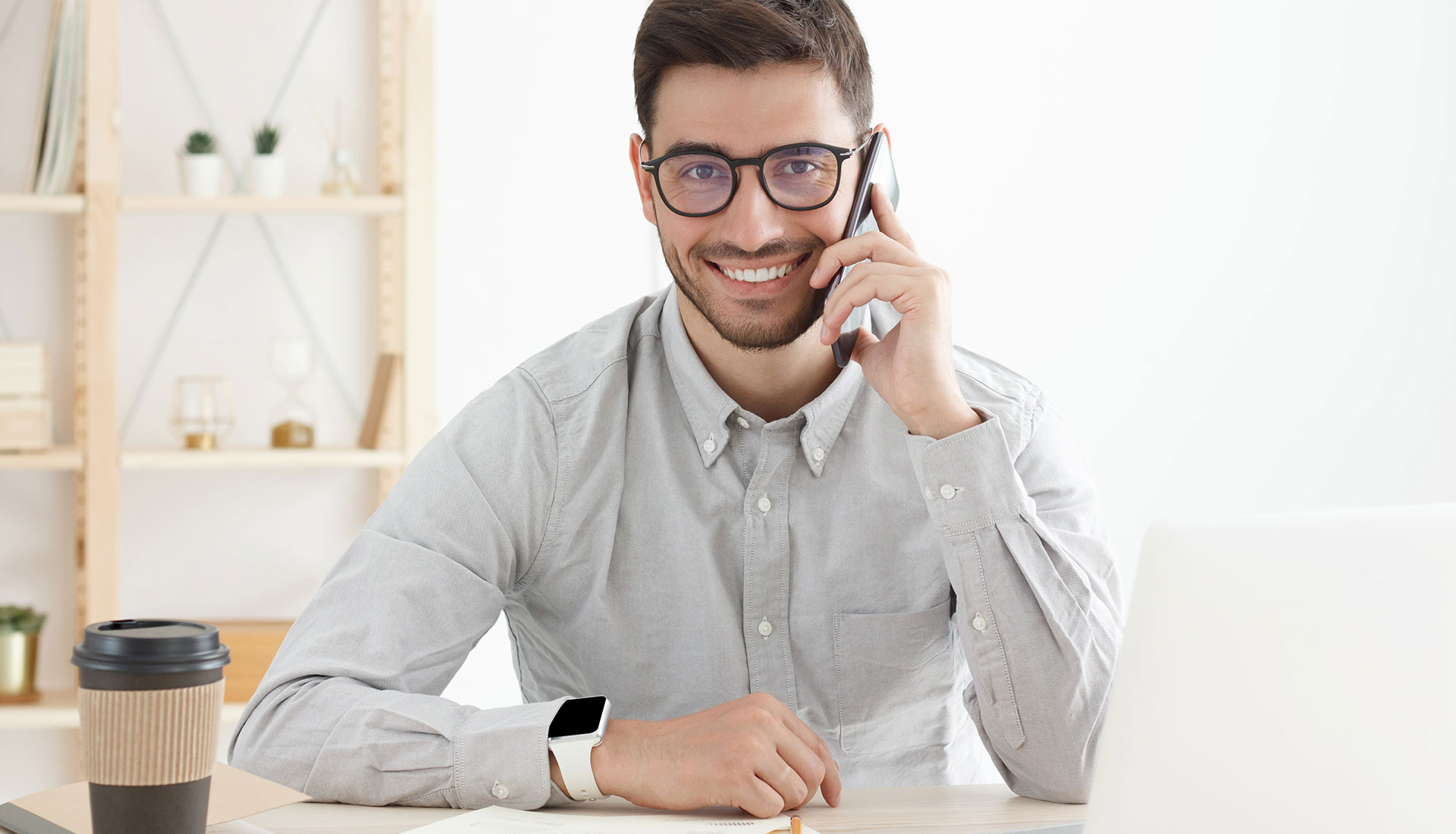 professioneller-berater-am-telefon Lächelnder Mann mit Brille sitzt am Schreibtisch, telefoniert und blickt auf einen Laptop. Neben ihm steht ein Coffee-to-go-Becher
